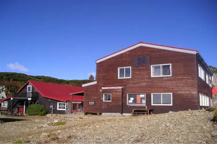 Wooden mountain huts with red roofs on rocky terrain under a clear blue sky.