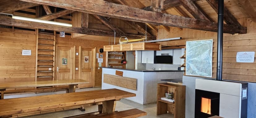 Interior of a rustic wooden mountain refuge dining area with long tables and a small kitchen counter.