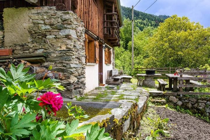 Stone and wood chalet exterior with pink peony, patio, and forested mountain backdrop in La Lechere.