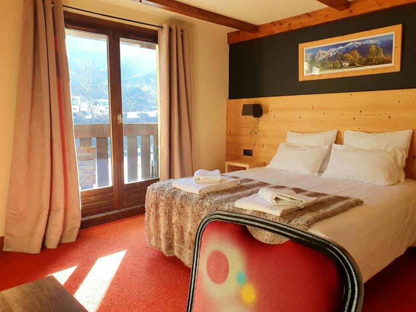 Chalet bedroom with wood paneling, white bedding, and balcony view of snowy mountains.