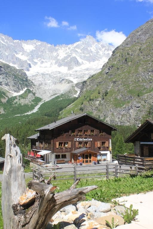Chalet L'Edelweiss in alpine valley with snow-capped mountains under blue sky