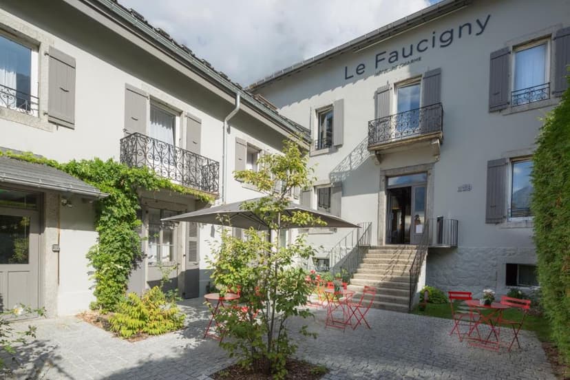 Hotel Le Faucigny exterior courtyard with cobblestones, red metal tables, and green foliage.