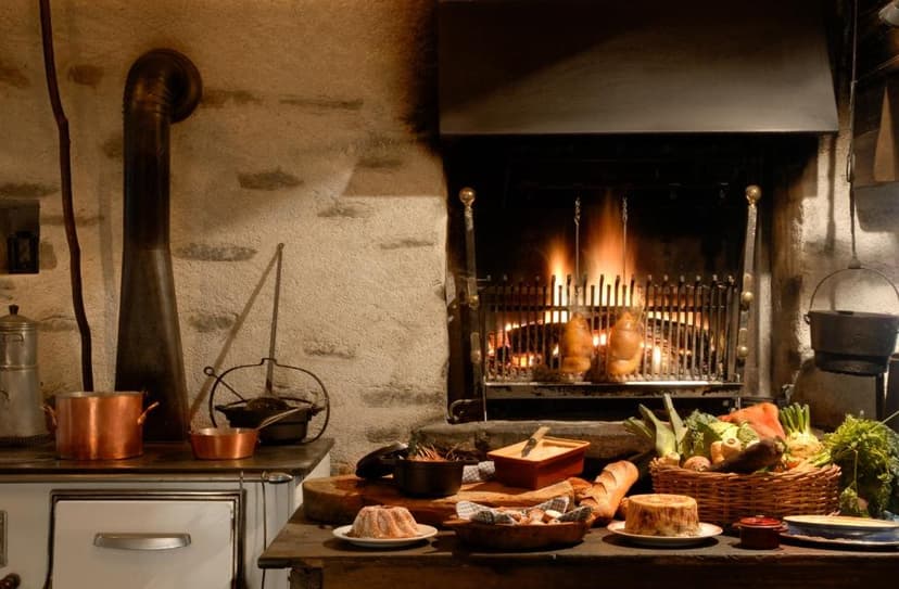 Rustic kitchen with food spread, copper pots, and meats roasting over a stone fireplace.