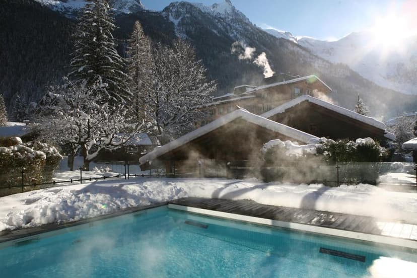 Outdoor heated pool steaming in snow with wooden chalets and sunny alpine mountains.