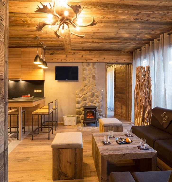 Rustic living room with wood paneling, stone fireplace, antler chandelier, and modern kitchen area.