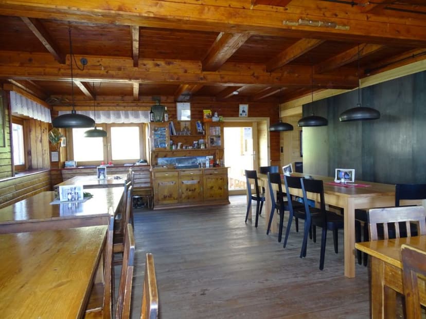 Rustic wooden interior dining room with long tables and black chairs, likely in a mountain hut.