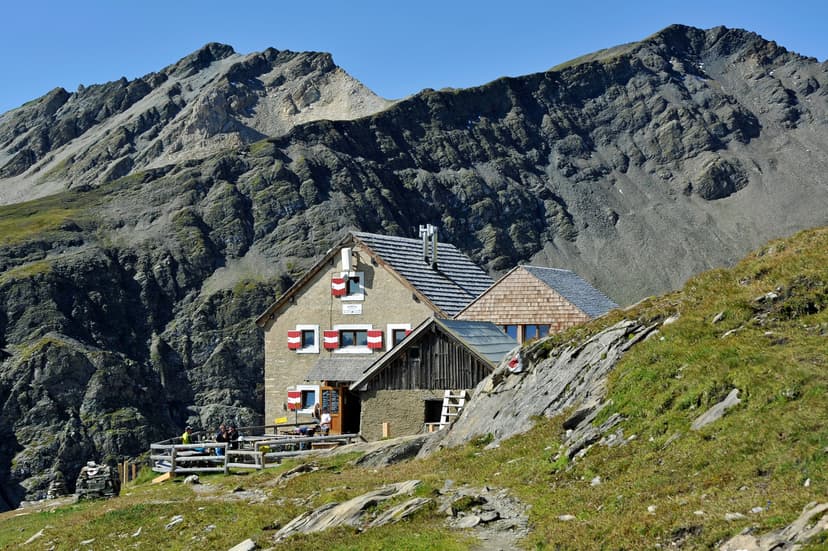 Salm Hutte mountain refuge with hikers resting below steep, rocky alpine slopes under a clear blue sky.