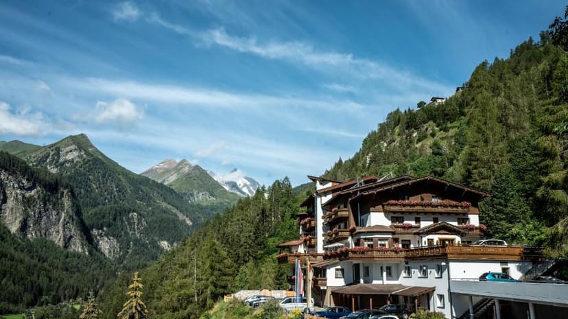 Hotel building nestled in a green alpine valley with forested mountains and snow-capped peaks.