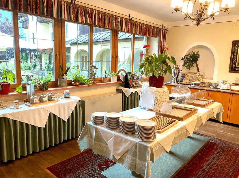 Hotel breakfast buffet setup with stacked plates, serving dishes, and plants by a large window overlooking a garden.