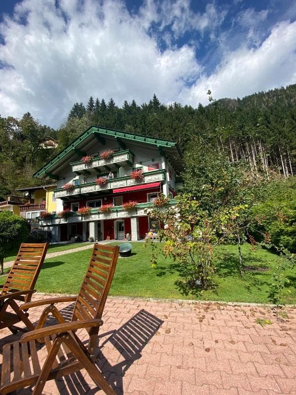 Wooden lounge chairs on patio overlooking traditional alpine apartment house and forested mountain.