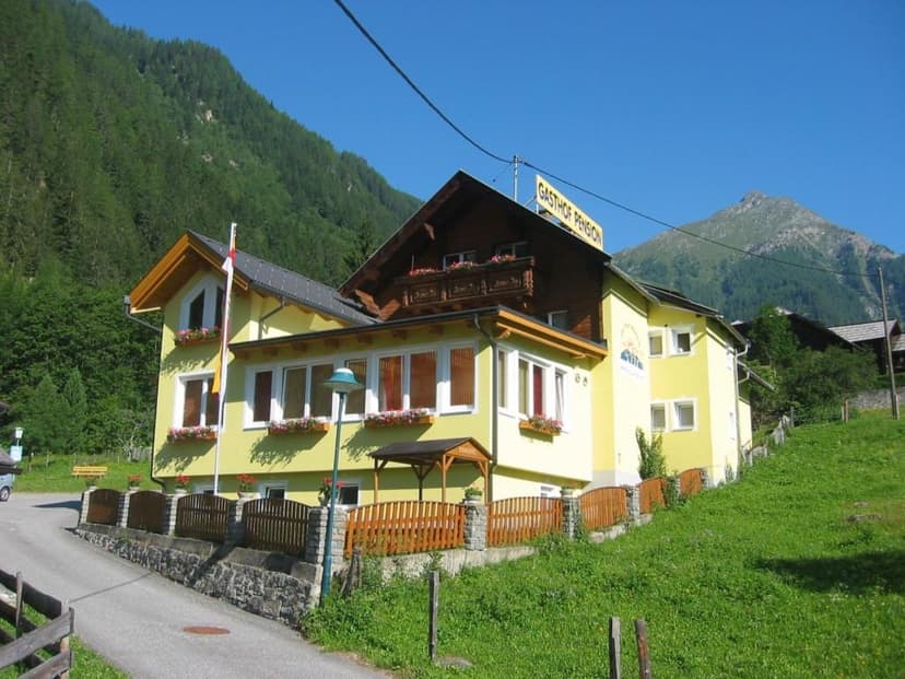 Yellow Gasthof Pension building on grassy slope with forested mountains under blue sky, Innerfragant.