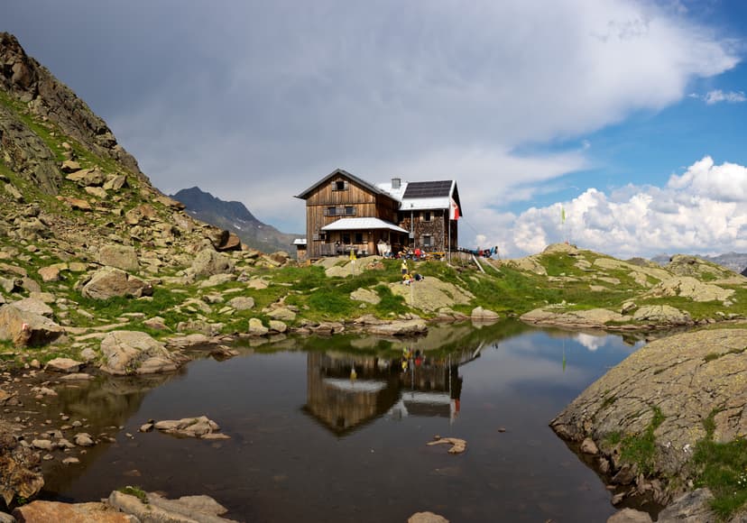 Wooden mountain hut reflected in a dark alpine pond with rocky terrain and mountains.