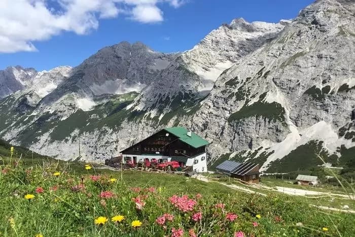Alpine hut with green roof nestled below rugged, rocky mountains and summer wildflowers.