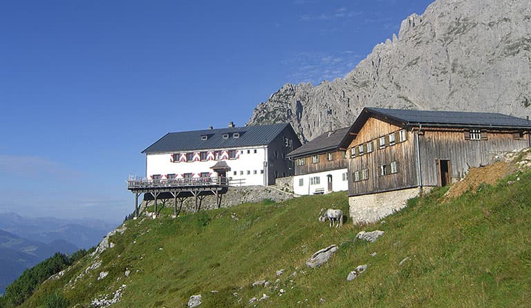 Gruttenhütte mountain hut with wooden buildings below a rocky peak on a grassy slope.