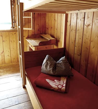 Bunk bed with red bedding and edelweiss blanket inside wooden paneled room at Reintalangerhütte.
