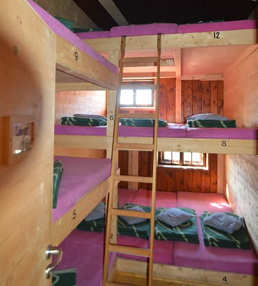 Wooden bunk beds with pink mattresses and green blankets inside a Knorrhütte mountain hut room.
