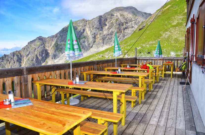 Outdoor wooden terrace with yellow tables overlooking steep green and rocky mountains.