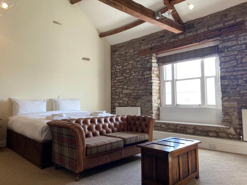Hotel room with exposed stone wall, wooden beams, bed, and leather Chesterfield sofa.