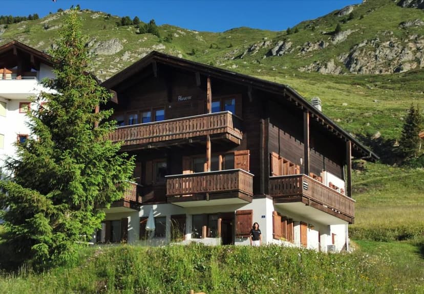 Wooden chalet in Riederalp against a green, rocky mountainside under a blue sky.