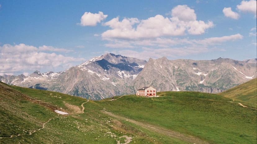Refuge Col de Balme hut on green hillside with snow-capped mountains under blue sky.