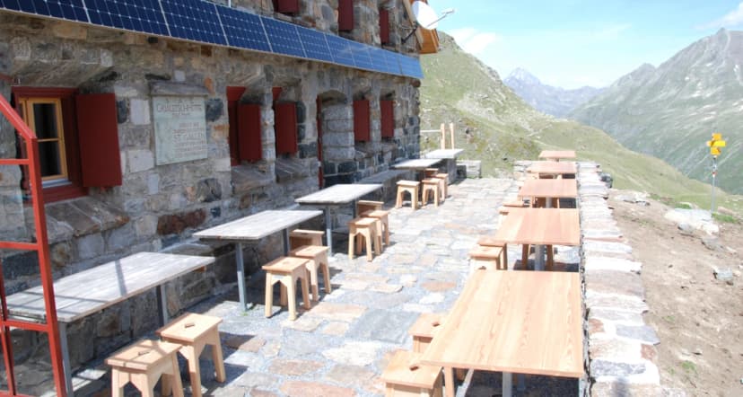Grialetsch Hut with solar panels, stone exterior, and outdoor wooden tables overlooking alpine mountains.