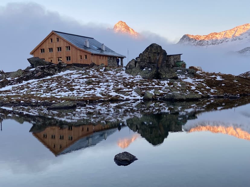 Wooden mountain hut reflected in still water near snow-dusted rocks at sunrise.