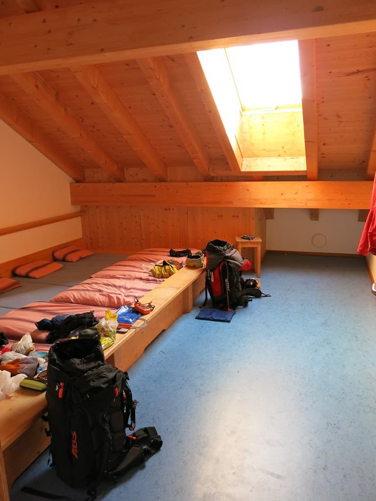 Backpacks and gear in a wooden dorm room with a bright skylight, likely a mountain hut.