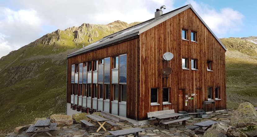 Wooden alpine hut with solar panels on windows, set against grassy mountainside with picnic tables.