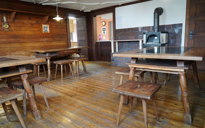 Rustic dining room with wood paneling, heavy wooden tables, stools, and a wood-burning stove.