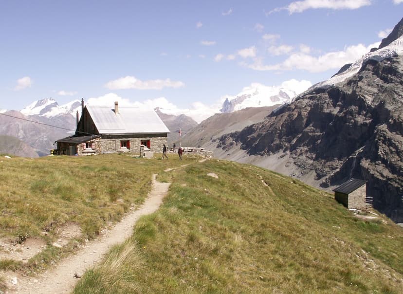 Stone mountain hut on grassy slope with hikers, snow-capped peaks in background