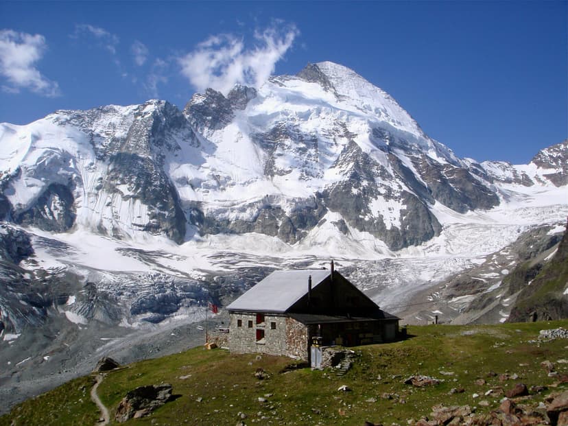 Stone mountain hut on grassy slope below massive snow-covered peaks and glacier.