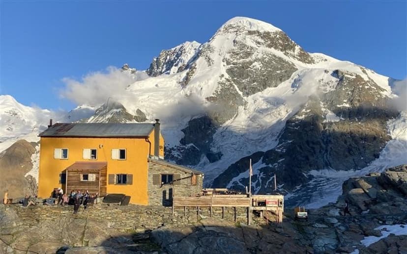 Gandegg Hut with hikers resting below a massive, snow-covered alpine peak.