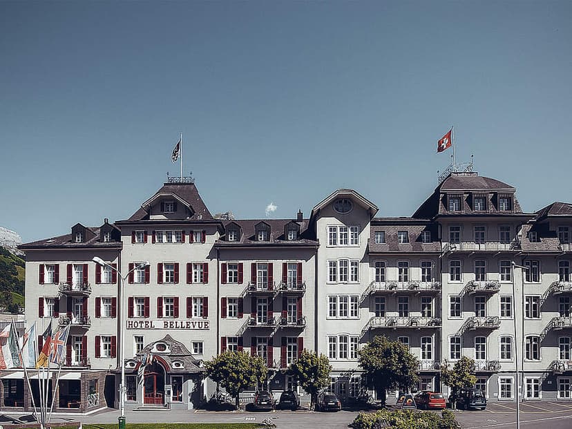Hotel Bellevue building with Swiss flag flying under a clear blue sky, mountains visible left.