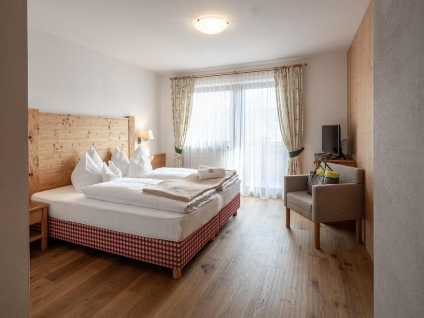 Hotel room with wooden headboard, white bedding, armchair, and sheer patterned curtains.