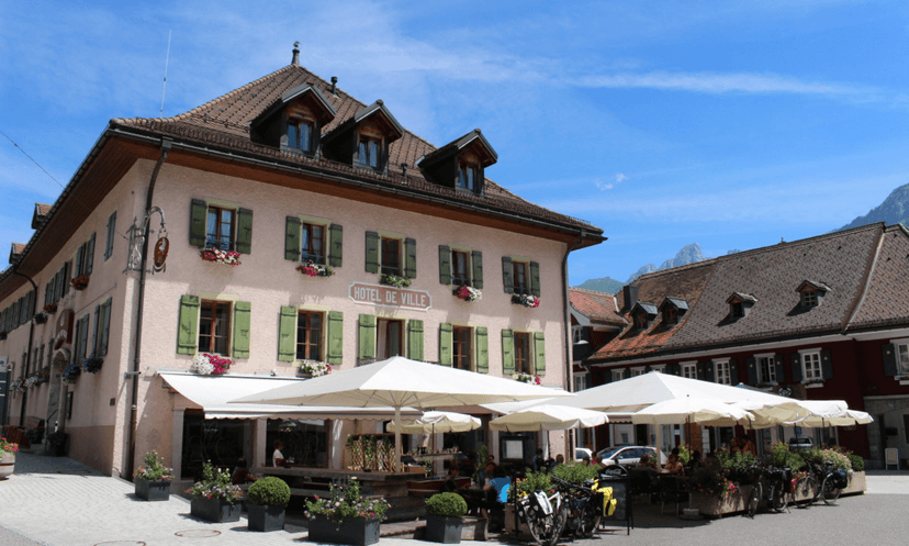 Hotel de Ville building with outdoor cafe seating under white umbrellas and mountains in background.