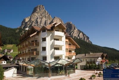 Hotel Col Alto building with balconies against a backdrop of rugged, forested mountains under a clear blue sky.