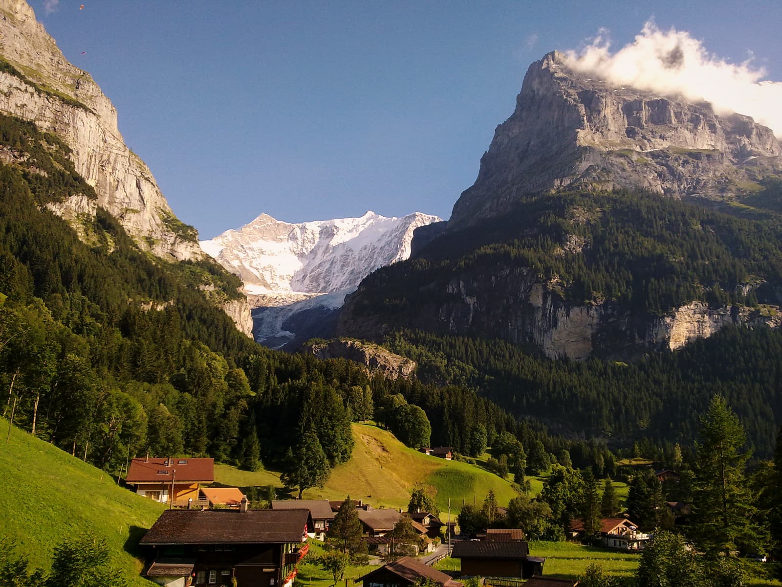 Alpine village nestled in valley with snow-capped mountains and glacier under blue sky.