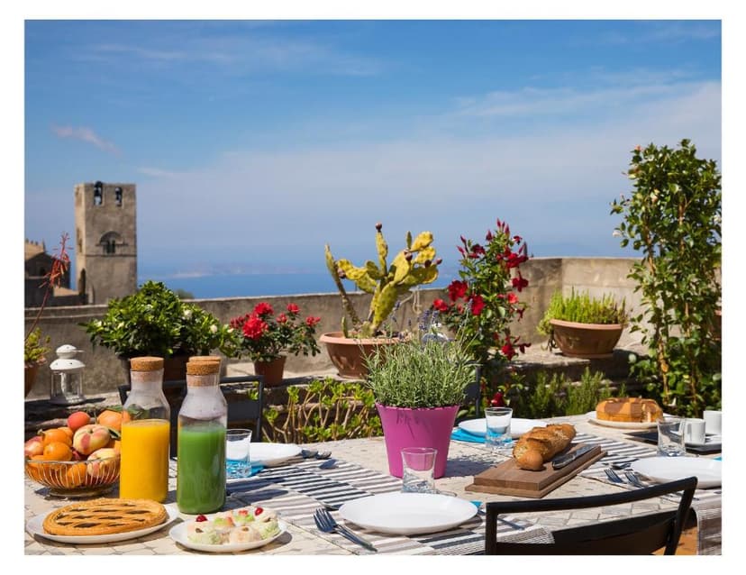 Outdoor breakfast terrace with fruit, juice, and bread overlooking the sea and an old stone tower.