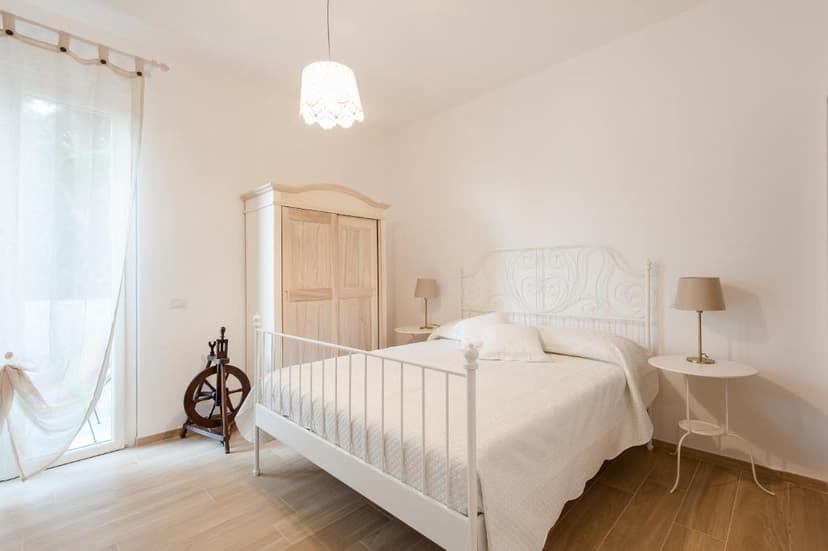 White bedroom with wrought iron bed, light wood wardrobe, and spinning wheel near sheer curtains.