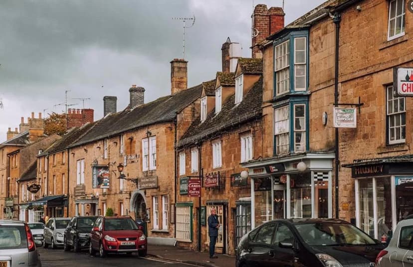 Row of historic stone buildings with shops and parked cars on a cloudy day.