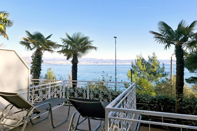 Balcony with lounge chairs overlooking sea, palm trees, and distant snow-capped mountains.