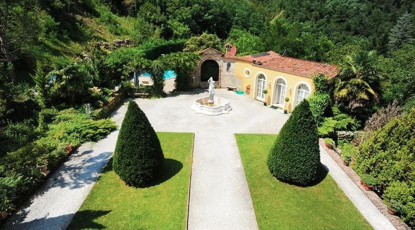 Villa courtyard with fountain, statue, manicured hedges, and lush green hillside.