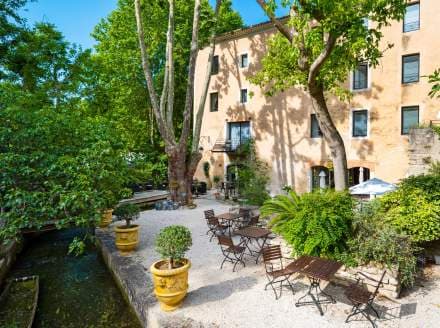 Outdoor dining area with tables near a stone building and shaded by large trees next to a canal.