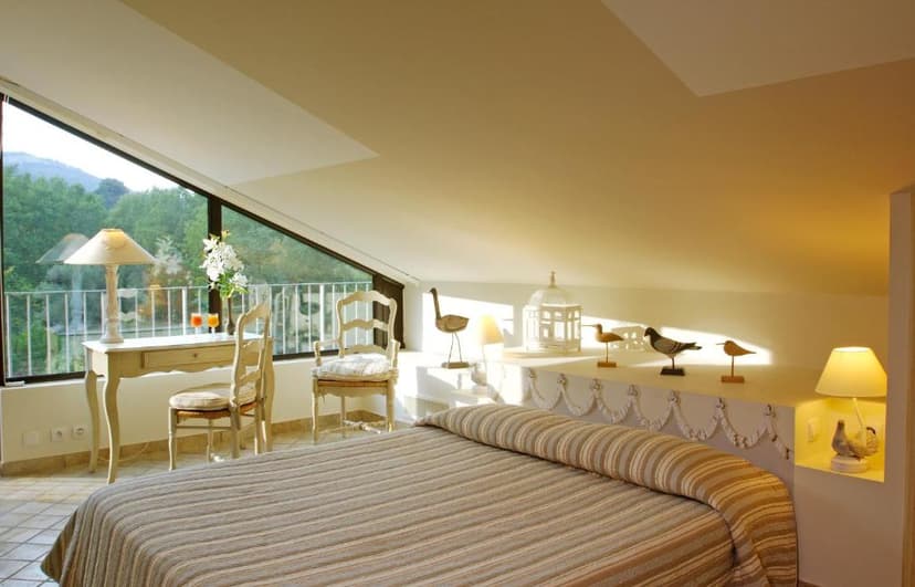 Attic bedroom with striped duvet, writing desk, and view of green mountains through slanted window.