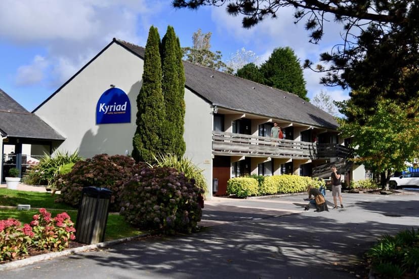 Kyriad Hotel building exterior with blue sign, surrounded by green trees and shrubs under a blue sky.