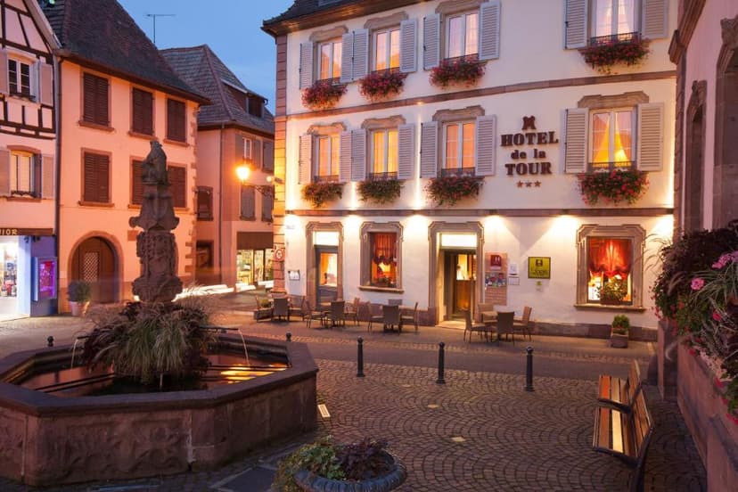 Hotel de la Tour with illuminated windows and outdoor seating facing a stone fountain square.