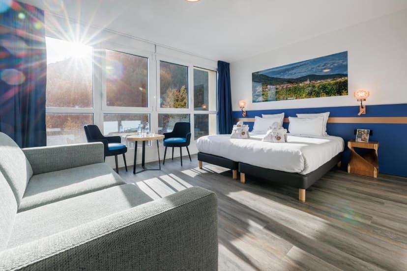 Bright hotel room with twin beds, grey sofa, and sunlit window view of trees, Husseren.