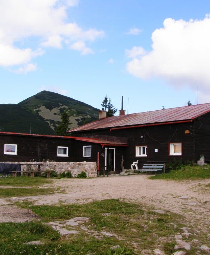 Mountain hut with dark wood exterior and red roof below a large green peak under a blue sky.