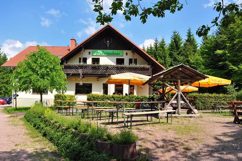 Hubertushaus restaurant with outdoor seating, yellow umbrellas, and surrounding forest on a sunny day.