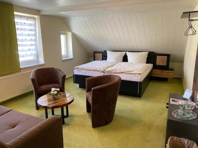 Hotel room with slanted ceiling, double bed, brown armchairs, and green carpet.